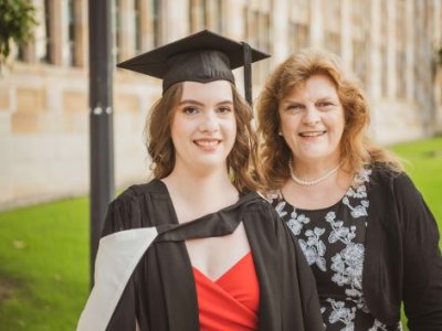 A young woman in graduation garb smiles next to an older woman in UQ's Great Court.